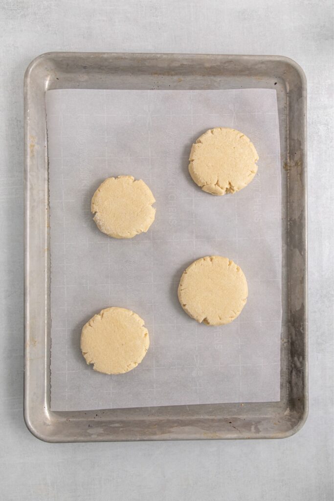 Four large disks of sugar cookie dough on a lined baking sheet.