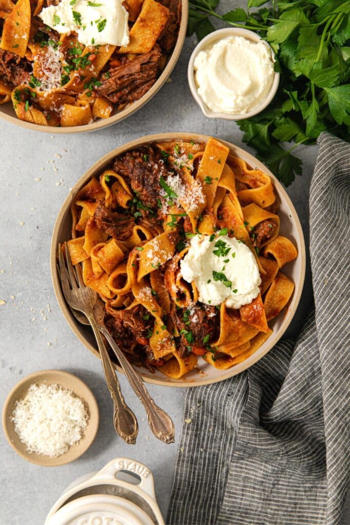 Two servings of beef short rib ragu with pappardelle garnished with a dollop of ricotta cheese, Parmesan, and fresh Italian parsley.