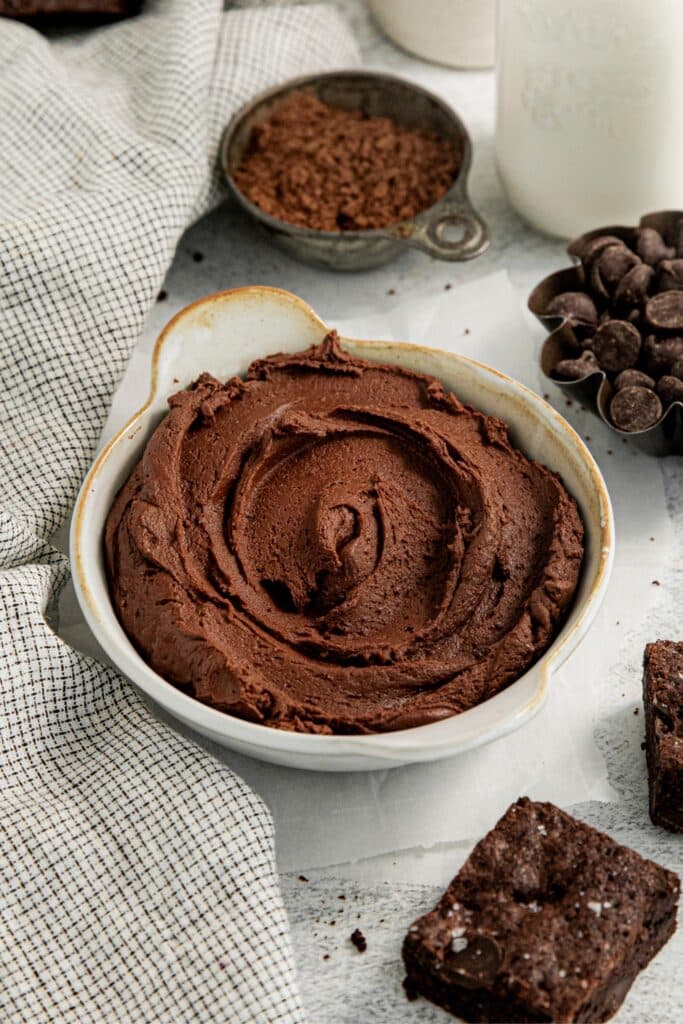 Thick, luscious brownie frosting in a small bowl next to some of the ingredients for making it.