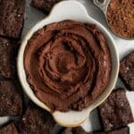 Brownie frosting in a small bowl that's surrounded by squares of brownies.