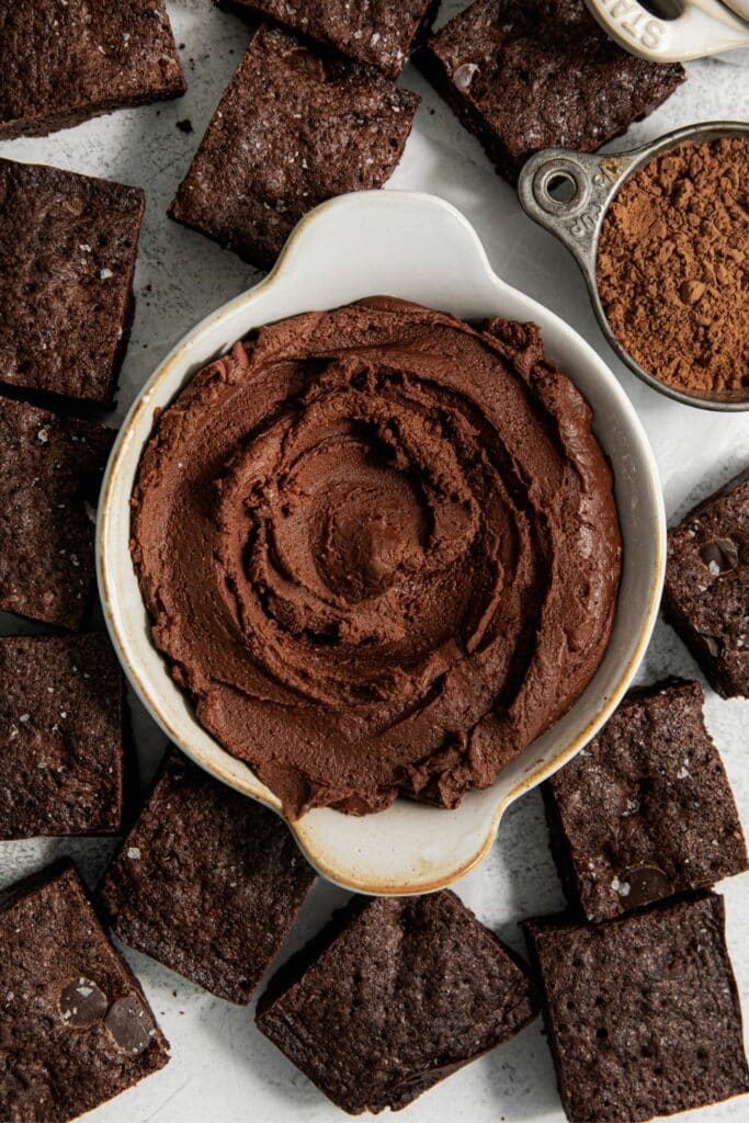 Brownie frosting in a small bowl that's surrounded by squares of brownies.