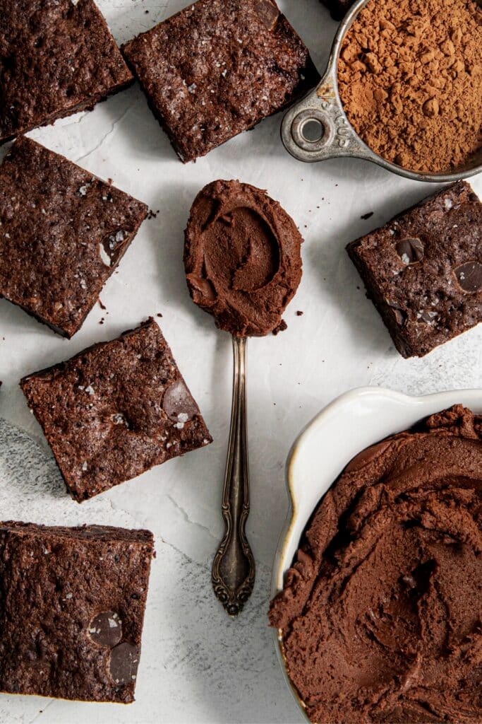 Brownie frosting in a bowl and on a spoon next to squares of dark chocolate brownies.
