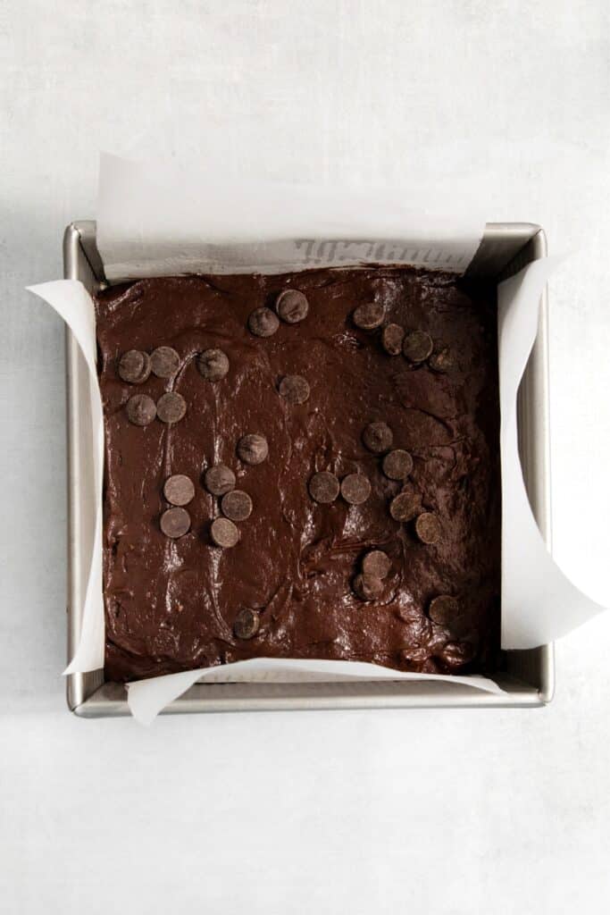Brownie batter with chocolate chips on top in a parchment paper-lined square metal pan.